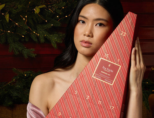 A woman holds a red Advent Calendar wrapped in diagonal striped paper with numbered windows. She is posed against a dark red wooden background with green pine branches and warm string lights.