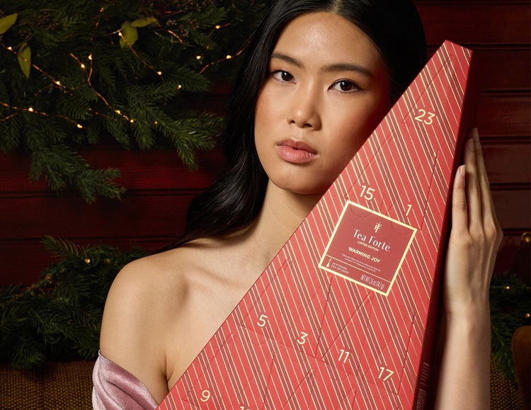 A woman holds a red Advent Calendar wrapped in diagonal striped paper with numbered windows. She is posed against a dark red wooden background with green pine branches and warm string lights.