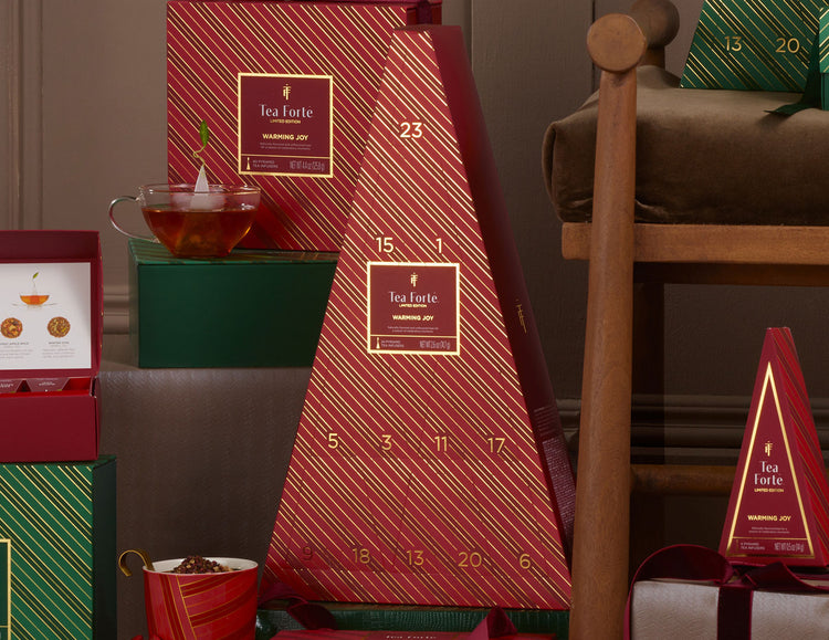 A red Advent Calendar with gold diagonal stripes and numbered compartments, displayed with matching gift boxes and a glass of tea in a styled indoor setting.