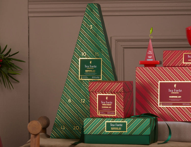 Tea Forté Herbal Advent Calendar of 24 pyramid infusers arranged on a bench against a brown paneled wall. The grouping includes several red striped boxes, and a green-and-red gift box tied with a ribbon.