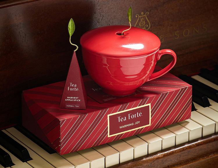 A red ceramic teacup with a matching lid sits on a red striped Tea Forte Petite Presentation Box, styled on piano keys with a tea bag tag visible.