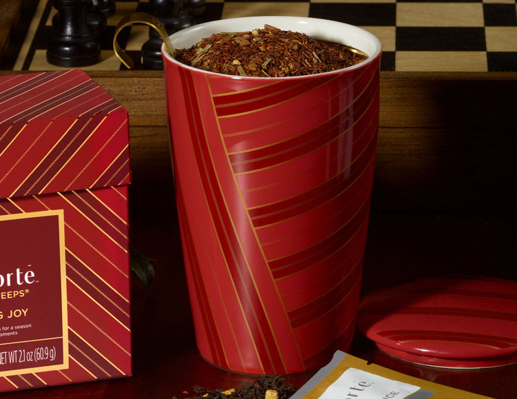 A red ceramic KATI Steeping Cup with gold diagonal stripes and a white interior, filled with a dark tea blend, shown beside its matching red box with gold stripes. A chessboard is blurred in the background.