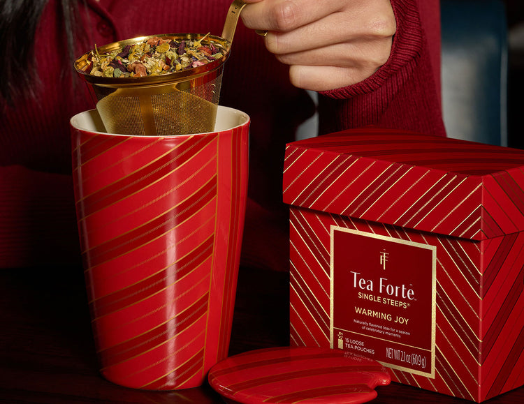 Woman in a red sweater pulls a loose-leaf tea gold strainer from a Warming Joy KATI Steeping Cup with diagonal red stripe patterns. A Warming Joy Single Steeps Sampler box sits beside the mug and it's lid on a dark table.