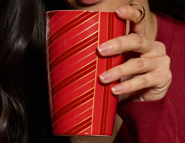 A close-up of a person holding a red KATI Steeping Cup Warming Joy with gold diagonal stripes. The person has long dark hair, is wearing a burgundy sweater, and a gold ring on their finger.