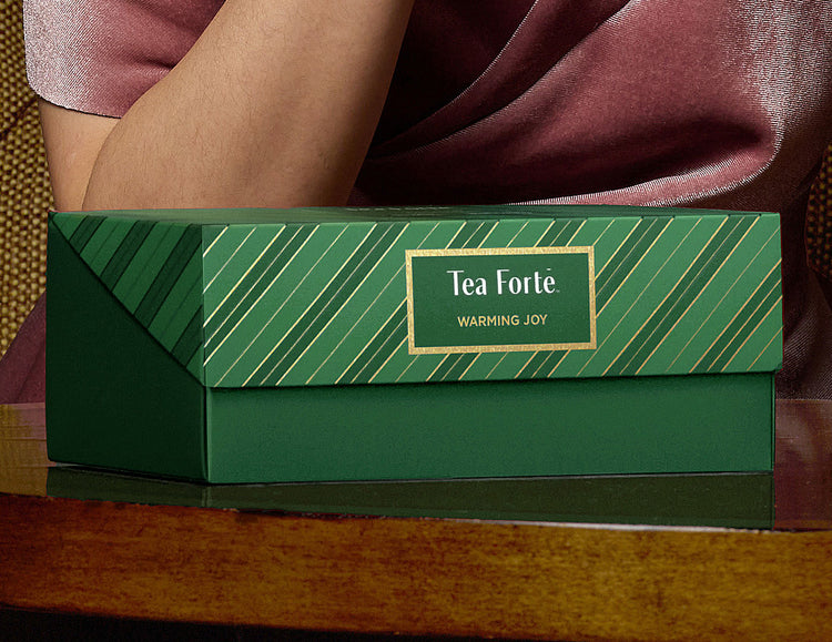A green striped Warming Joy Herbal Presentation Box 2026 of 20 pyrmid tea infusers, closed, placed on a wooden surface with a person in a mauve dress partially visible behind it.
