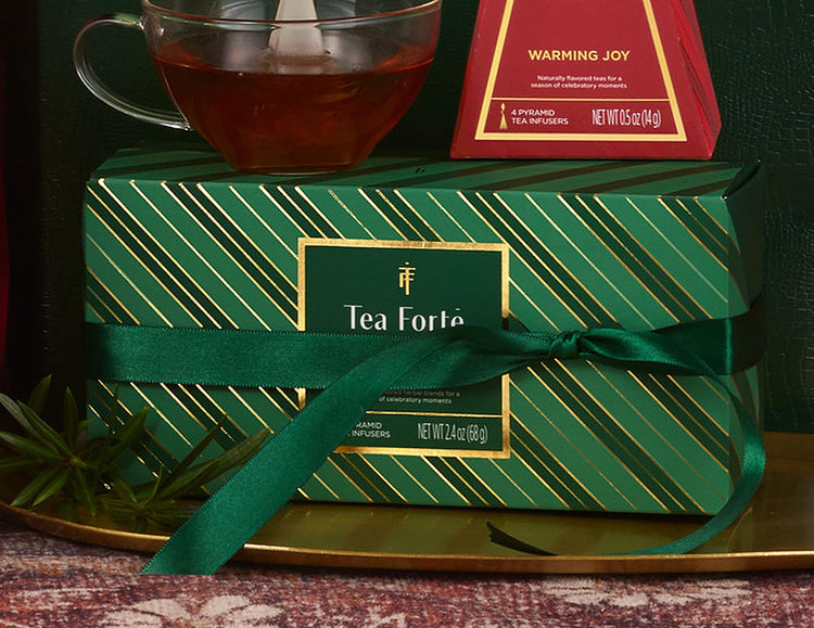 A Warming Joy Herbal Presentation Box of 20 pyrmid tea infusers, set on a gold tray, with a green ribbon tied around it, a glass cup of amber tea above, and a Warming Joy Petite Tea Tree behind it.