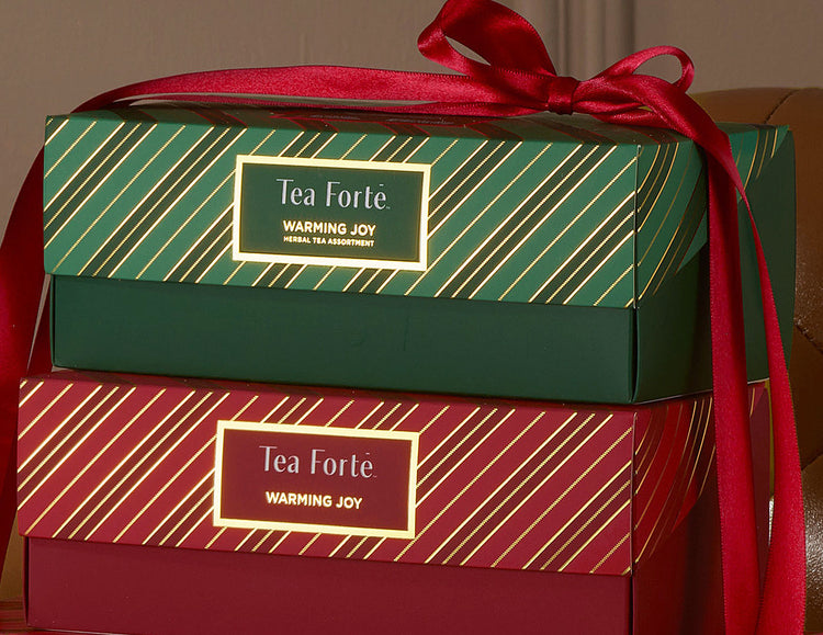 Two stacked Warming Joy Presentation Boxes with a red satin ribbon tied around both. The top box is dark green with gold diagonal stripes, and the bottom box is deep red with gold diagonal stripes.