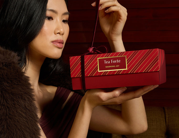 A woman with a fur shawl unties the  red ribbon around a Presentation Box Warming Joy, of 20 tea infusers, against a dark red background.