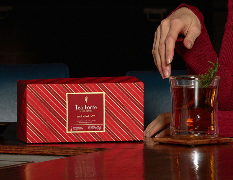 Presentation Box Warming Joy, of 20 tea infusers, closed, with diagonal gold stripes, placed on a dark wooden table beside a glass of iced tea with a rosemary sprig. A person in a red sweater is partially visible reaching toward the drink.