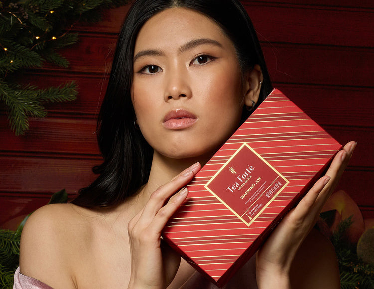A woman holds a Petite Presentation Box Warming Joy against a dark red holiday-themed background with pine branches.