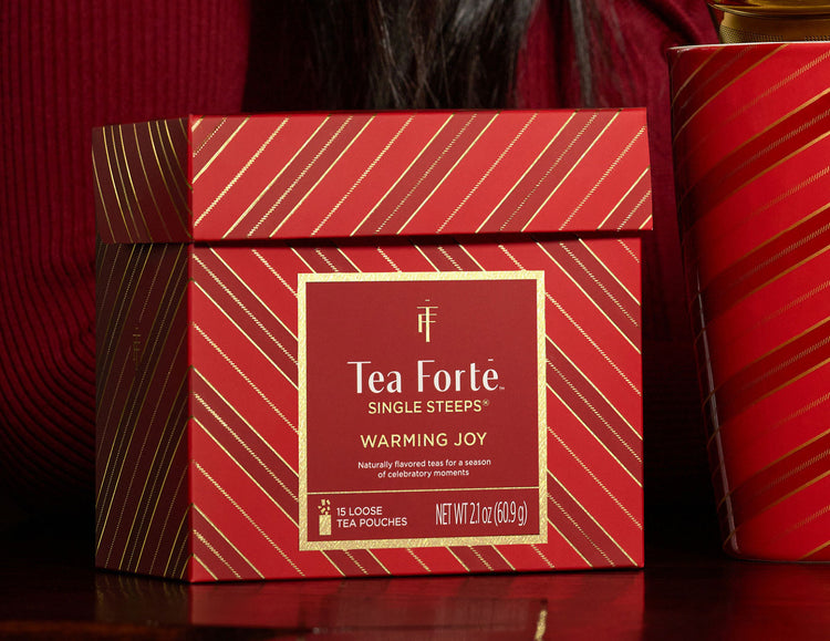 Warming Joy Single Steeps Sampler of 15 loose tea pouches, shown on a dark reflective surface with other cups and packages in the background