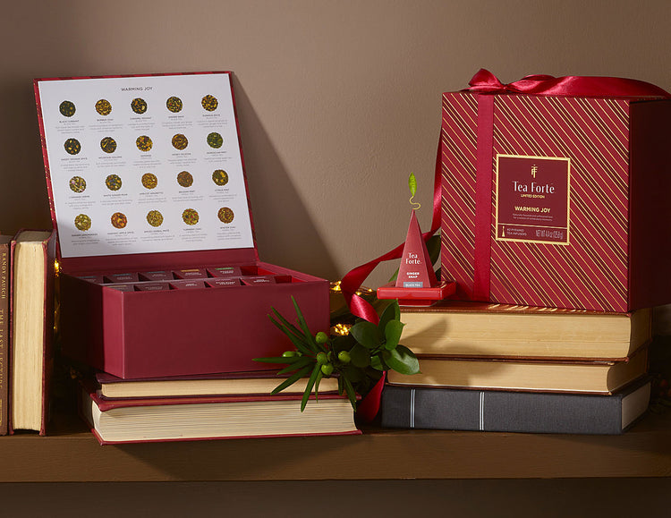 A red Tea Chest Warming Joy of 40 teas, open and closed, displayed on stacked books against a warm brown background and green foliage.