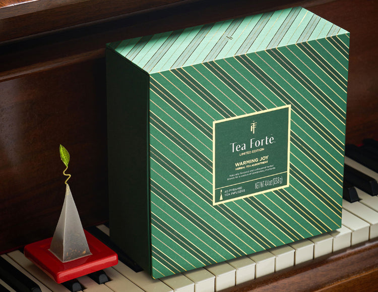 Warming Joy Herbal Tea Chest closed, displayed on a piano next to a red Tea Tray with a tea infuser on top.