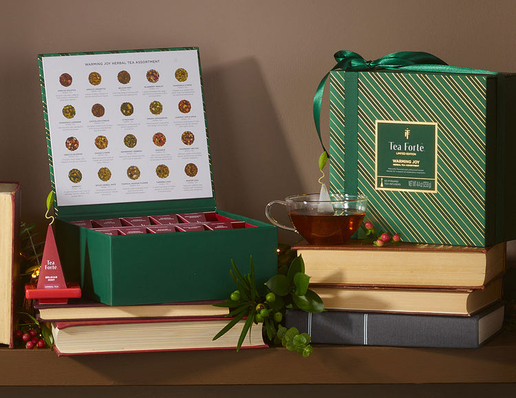 Tea Chest Herbal Warming Joy tied with a ribbon, displayed on stacked books with a glass teacup and infuser.