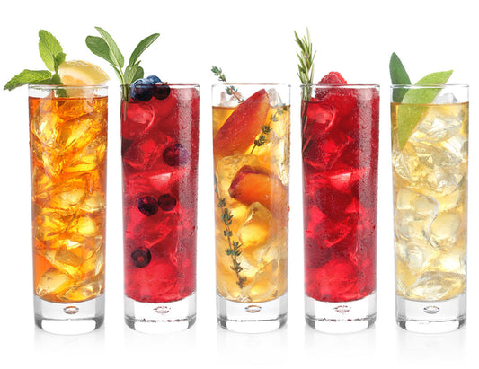 Five tall clear glasses filled with iced tea and garnishes, arranged in a row on a white background. The drinks show orange, red, and pale yellow tones with visible ice cubes, mint, blueberries, peach slices, rosemary, and lemon.