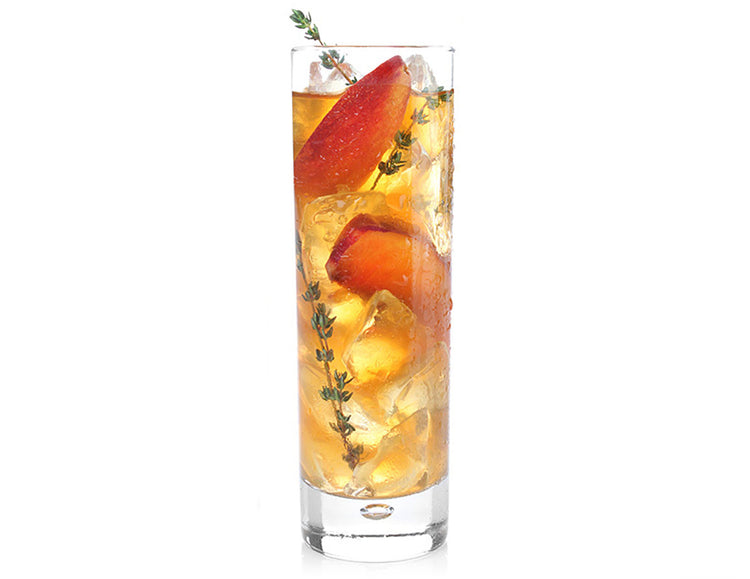 A clear highball glass filled with an Green Mango Peach iced tea, ice cubes, peach slices, and sprigs of fresh thyme, isolated on a white background.