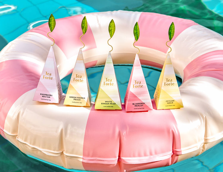 Five pyramid infusers of iced tea, arranged on a pink and white striped inflatable pool float over turquoise water. The tea bags feature different flavor labels including Raspberry Nectar, Green Mango Peach, White Ginger Pear, Blueberry Merlot, and Ceylon Gold.