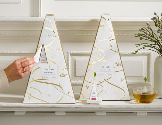 Winter Chalet Advents, one with doors closed, the other being opened with a female hand, on a white shelf with glass teacup of tea