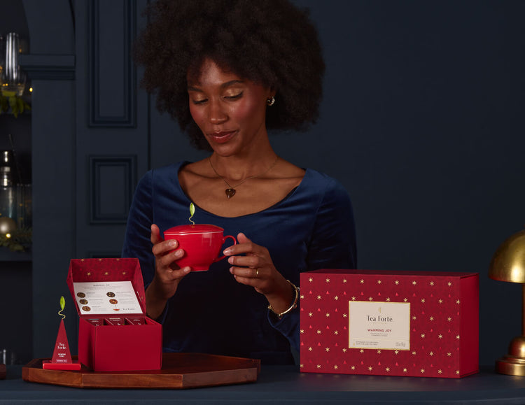 Woman holding a red mug of the Warming Joy Gift Set, surrounded by tea packaging on a dark blue background